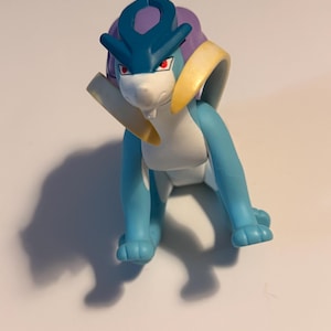 Puede incluir: Una figura de Suicune, un personaje de Pokémon, se muestra en una pose frontal. La figura es principalmente azul con detalles blancos, una melena púrpura y detalles amarillos y blancos. Tiene ojos rojos y está sobre un fondo blanco.