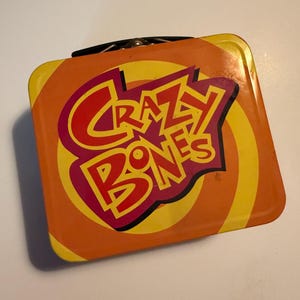 May include: A square, orange metal lunchbox with the words "Crazy Bones" in red and yellow. The lunchbox has a black handle and a spiral design on the front.