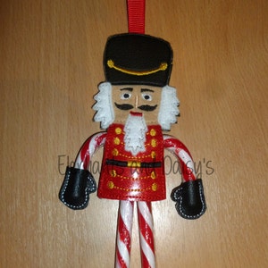 May include: A felt nutcracker ornament with a candy cane body, black boots, and a red and gold uniform. The ornament is hanging from a red ribbon.