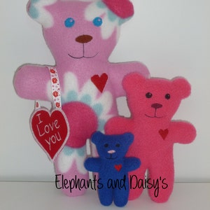 May include: Three plush teddy bears, one pink with white and blue floral print, one pink, and one blue. The pink bear with the floral print has a heart-shaped tag that says "I Love You".