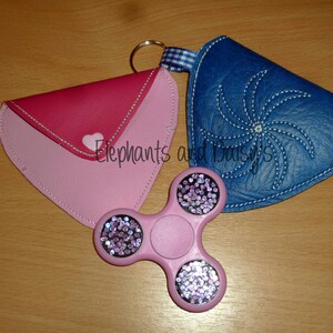 May include: Two pink and blue fabric coin purses with a pink and purple fidget spinner in the center. The purses have a heart and a swirl design.