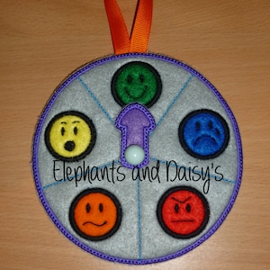 May include: A gray felt wheel with a purple border and a blue and white arrow in the center. The wheel has six different colored felt circles with different facial expressions on them. The text "Elephants and Daisy's" is printed on the wheel.