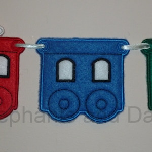 May include: A red, blue, and green felt train banner with three cars. The first car is a red engine with a white smoke plume. The second car is blue with two windows. The third car is green.