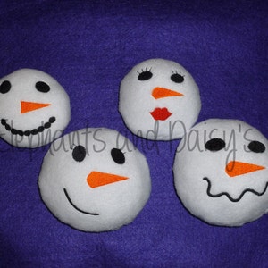 May include: Four white felt snowman ornaments with black eyes, orange noses, and embroidered mouths. The snowman on the left has a black smile, the snowman in the center has a black smile and eyelashes, the snowman on the right has a black frown, and the snowman in the back has a red mouth.