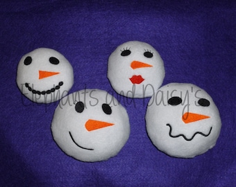 May include: Four white felt snowman ornaments with black eyes, orange noses, and embroidered mouths. The snowman on the left has a black smile, the snowman in the center has a black smile and eyelashes, the snowman on the right has a black frown, and the snowman in the back has a red mouth.