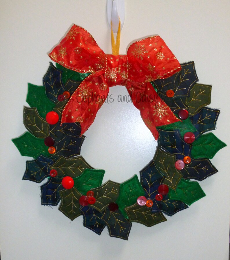 May include: A Christmas wreath made of green and blue felt leaves with red and orange buttons and a red bow with gold glitter.