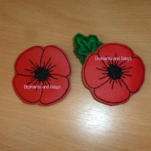 May include: Two red felt poppy flowers with black embroidered centers and green felt leaves. The flowers have the text "Elephants and Daisys" embroidered on them.