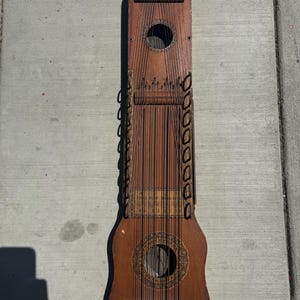 May include: A vintage, wooden musical instrument with numerous strings. The instrument has a dark brown finish, with decorative circular sound holes and intricate stringing patterns. The instrument's design suggests it may be a type of zither or similar stringed instrument.