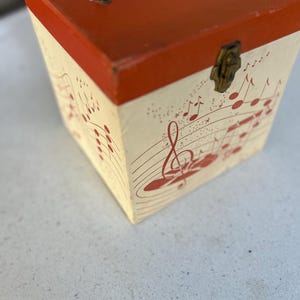 May include: A vintage square record case with a red top and a cream-colored body. The case features a clear plastic handle and a brass clasp. The body is decorated with red musical notes and staff lines, creating a musical theme.