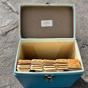May include: An open, vintage blue record storage box with a brass clasp. The interior features labeled dividers and a label that reads "Capitol". The box is sitting on a gray surface.