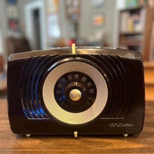 May include: A vintage black RCA Victor radio with a circular dial and gold accents. The radio features a tuning dial with numbers and the RCA Victor logo. The radio is set on a wooden surface.