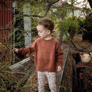 Puede incluir: Un niño pequeño con un suéter de punto color óxido y leggings blancos con lunares negros. El niño está de pie en una plataforma de madera con un fondo verde frondoso.