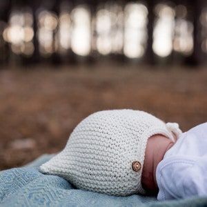K&ouml;nnte beinhalten: Eine wei&szlig;e gestrickte Babyhaube mit einem Holzknopf an der Seite. Die Haube liegt auf einer blau-gr&uuml;nen gemusterten Decke.