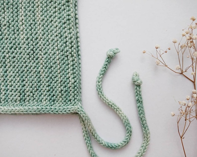 K&ouml;nnte beinhalten: Eine gr&uuml;ne Strickm&uuml;tze mit einem wei&szlig;en und gr&uuml;nen Ombre-Effekt. Die M&uuml;tze hat zwei lange B&auml;nder, die in Knoten gebunden sind.