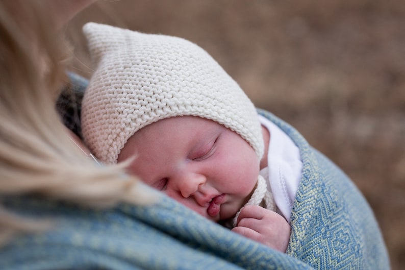 K&ouml;nnte beinhalten: Ein Baby in einer wei&szlig;en Strickm&uuml;tze schl&auml;ft friedlich in einem blauen und gr&uuml;nen gemusterten Babytragetuch.