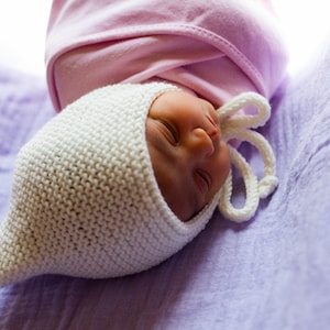 K&ouml;nnte beinhalten: Ein neugeborenes Baby, das eine wei&szlig;e H&auml;kelm&uuml;tze mit einer rosa Schleife oben tr&auml;gt, schl&auml;ft auf einer lila Decke.