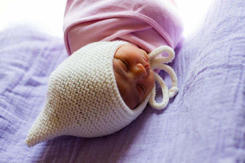 K&ouml;nnte beinhalten: Ein neugeborenes Baby, das eine wei&szlig;e H&auml;kelm&uuml;tze mit einer rosa Schleife oben tr&auml;gt, schl&auml;ft auf einer lila Decke.