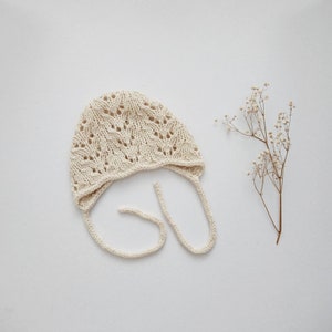 May include: A white crocheted baby bonnet with a delicate lace pattern and ties. The bonnet is on a white background.