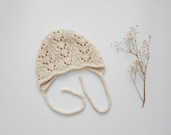 May include: A white crocheted baby bonnet with a delicate lace pattern and ties. The bonnet is on a white background.