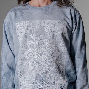 Pode incluir: Camisola azul clara com um design de mandala floral branco. A parte superior de manga comprida tem um efeito tie-dye e gola redonda. O gr&aacute;fico est&aacute; centrado na frente da pe&ccedil;a.