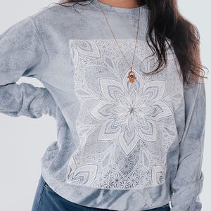 Pode incluir: Uma camisola cinza claro com um desenho de mandala branco. A parte superior de manga comprida &eacute; combinada com cal&ccedil;as de carpinteiro de ganga azul. Um colar de ouro com um pingente &eacute; vis&iacute;vel.
