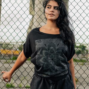 May include: Black t-shirt with a large, detailed gray mandala design on the front. The shirt has short sleeves and a scoop neck. The person is wearing black cargo pants. The background features a chain-link fence.