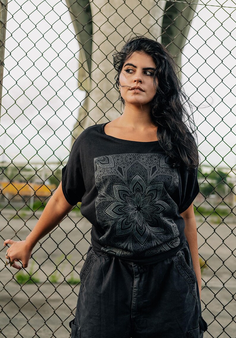 May include: Black t-shirt with a large, detailed gray mandala design on the front. The shirt has short sleeves and a scoop neck. The person is wearing black cargo pants. The background features a chain-link fence.