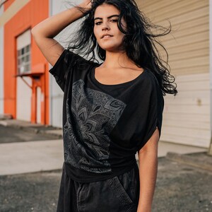 May include: Black scoop-neck t-shirt with a gray floral design. The shirt has short sleeves and is paired with black pants. The model is standing in front of a building with orange and white walls.