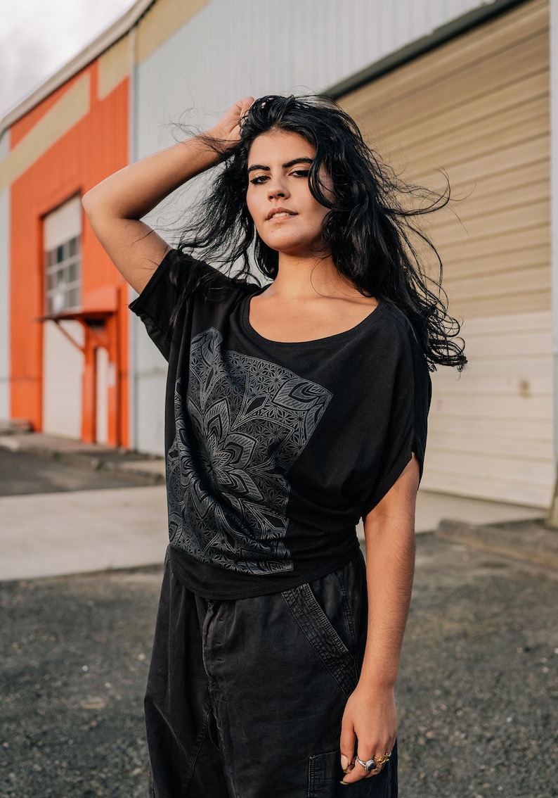 May include: Black scoop-neck t-shirt with a gray floral design. The shirt has short sleeves and is paired with black pants. The model is standing in front of a building with orange and white walls.