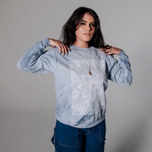 Pode incluir: Camisola azul clara de manga comprida com um desenho floral branco na frente. A camisola tem um efeito tie-dye nas mangas e &eacute; combinada com macac&atilde;o de ganga azul escuro. Um colar de ouro tamb&eacute;m &eacute; vis&iacute;vel.