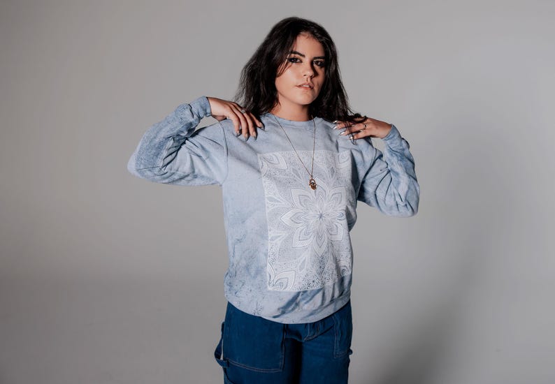 Pode incluir: Camisola azul clara de manga comprida com um desenho floral branco na frente. A camisola tem um efeito tie-dye nas mangas e &eacute; combinada com macac&atilde;o de ganga azul escuro. Um colar de ouro tamb&eacute;m &eacute; vis&iacute;vel.