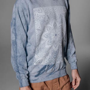 Pode incluir: Camisola azul tie-dye com um design floral de mandala branco na frente. A parte superior de manga comprida tem decote redondo. O modelo est&aacute; a usar cal&ccedil;as castanhas. O material parece macio.