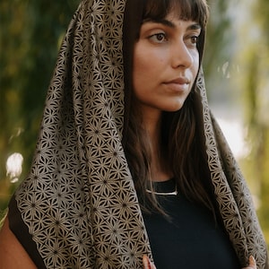 Peut inclure: Un foulard marron fonc&eacute; orn&eacute; d'un motif g&eacute;om&eacute;trique r&eacute;p&eacute;titif en forme d'&eacute;toile dans une teinte beige plus claire. Le tissu se drape sur la t&ecirc;te et les &eacute;paules, mettant en valeur un design sym&eacute;trique d&eacute;taill&eacute; couvrant la surface du textile.