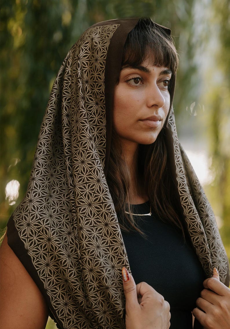 Peut inclure: Un foulard marron fonc&eacute; orn&eacute; d'un motif g&eacute;om&eacute;trique r&eacute;p&eacute;titif en forme d'&eacute;toile dans une teinte beige plus claire. Le tissu se drape sur la t&ecirc;te et les &eacute;paules, mettant en valeur un design sym&eacute;trique d&eacute;taill&eacute; couvrant la surface du textile.