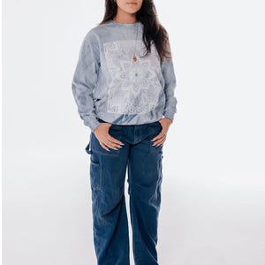 Pode incluir: Uma pessoa vestindo um moletom azul claro tie-dye com um design floral branco e jeans de carpinteiro azul escuro. A pessoa tamb&eacute;m est&aacute; usando um colar e sapatos de plataforma. O fundo &eacute; branco.