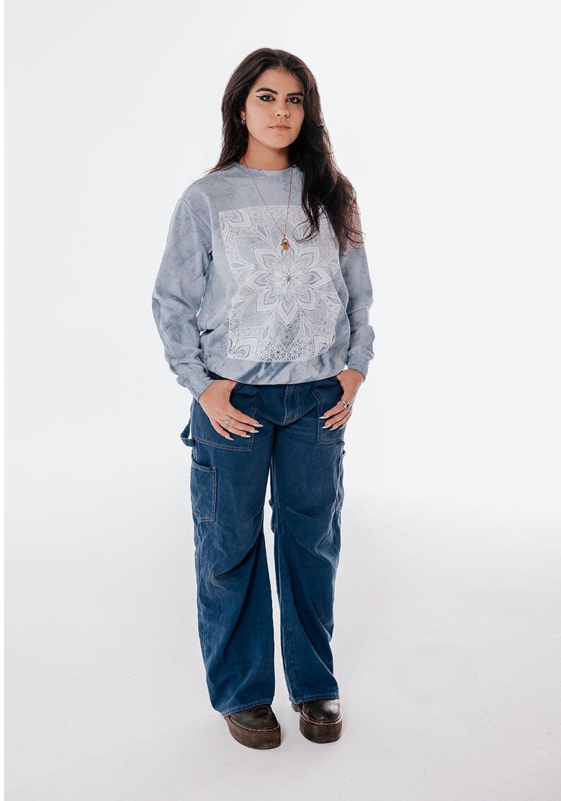 Pode incluir: Uma pessoa vestindo um moletom azul claro tie-dye com um design floral branco e jeans de carpinteiro azul escuro. A pessoa tamb&eacute;m est&aacute; usando um colar e sapatos de plataforma. O fundo &eacute; branco.