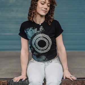 May include: A woman with curly brown hair is wearing a black t-shirt with a white and gray circular design. The design features a repeating pattern of letters and symbols. She is also wearing light blue jeans and has a silver necklace with a small pendant.