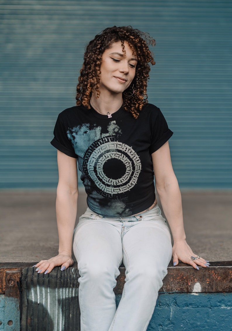 May include: A woman with curly brown hair is wearing a black t-shirt with a white and gray circular design. The design features a repeating pattern of letters and symbols. She is also wearing light blue jeans and has a silver necklace with a small pendant.