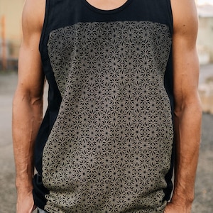 Puede incluir: Camiseta sin mangas negra con un patrón geométrico dorado en el pecho. La camiseta sin mangas tiene cuello redondo y está hecha de un material suave. El diseño presenta un motivo floral repetitivo. El modelo lleva pantalones grises.