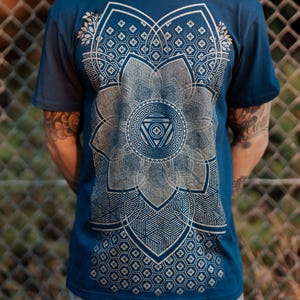 May include: A navy blue t-shirt featuring a large, intricate geometric design in shades of gray and white. The design is centered on the chest and resembles a stylized flower or mandala. The shirt has short sleeves.
