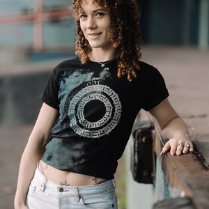 May include: A woman with curly brown hair is wearing a black t-shirt with a white graphic design of a circle with a pattern inside. She is also wearing light blue jeans and has a small tattoo on her hip.