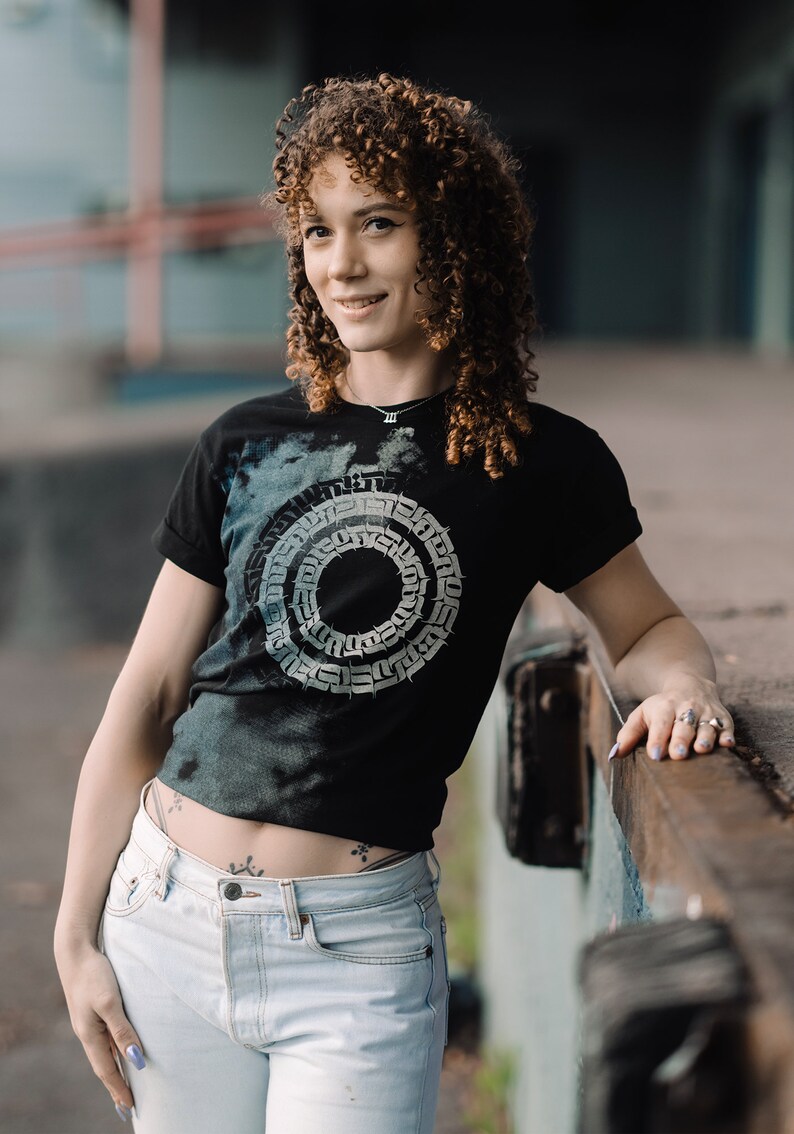 May include: A woman with curly brown hair is wearing a black t-shirt with a white graphic design of a circle with a pattern inside. She is also wearing light blue jeans and has a small tattoo on her hip.
