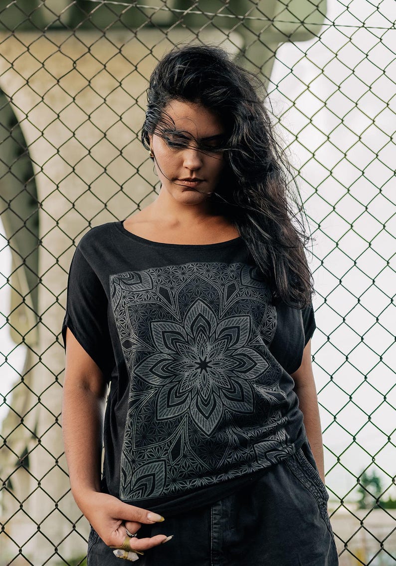 May include: Black t-shirt with a large, detailed gray mandala design on the front. The shirt has short sleeves and a scoop neck. The person is standing in front of a chain-link fence.