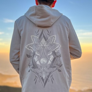 Puede incluir: Una sudadera con capucha gris claro con un gráfico de dos polillas en forma de diamante con una luna en el centro. Las polillas están delineadas en negro y tienen un relleno blanco. La sudadera tiene una capucha y una cremallera.