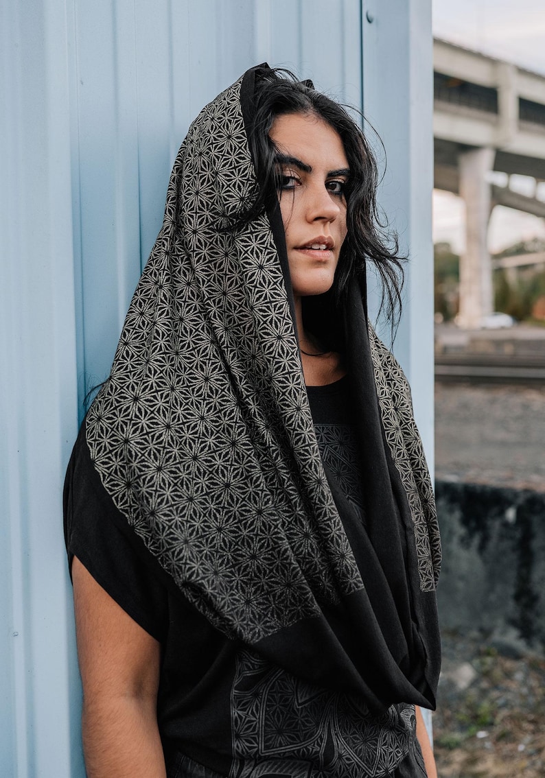 Peut inclure: Un capuchon noir avec un motif g&eacute;om&eacute;trique d'&eacute;toiles r&eacute;p&eacute;titives dans un ton dor&eacute; att&eacute;nu&eacute;. Le tissu se drape sur la t&ecirc;te et les &eacute;paules, mettant en valeur un design d&eacute;taill&eacute; et complexe qui couvre la capuche et descend sur le devant du v&ecirc;tement.
