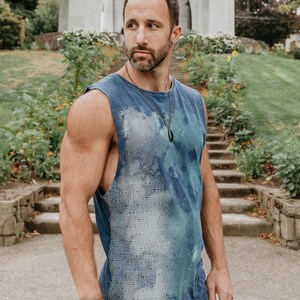May include: A man wearing a blue sleeveless shirt with a white geometric pattern. The shirt has a tie-dye effect and the man is wearing a black pendant necklace.
