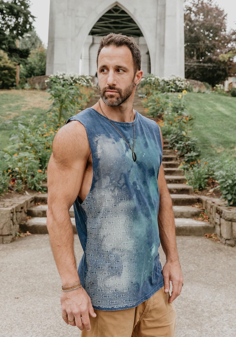May include: A man wearing a blue sleeveless shirt with a white geometric pattern. The shirt has a tie-dye effect and the man is wearing a black pendant necklace.