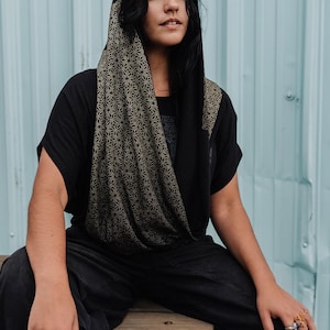 Peut inclure: Un haut &agrave; capuche noir avec un motif g&eacute;om&eacute;trique en forme d'&eacute;toile dor&eacute;e sur la capuche et l'encolure b&eacute;nitier. Le v&ecirc;tement a des manches courtes et est associ&eacute; &agrave; un pantalon noir. La capuche est rabattue sur la t&ecirc;te, r&eacute;v&eacute;lant le motif r&eacute;p&eacute;titif.