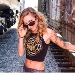 May include: A woman wearing a black crop top with a gold mandala and lotus flower design. She is wearing sunglasses and has a tattoo on her arm.