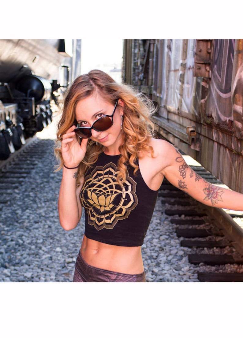 May include: A woman wearing a black crop top with a gold mandala and lotus flower design. She is wearing sunglasses and has a tattoo on her arm.